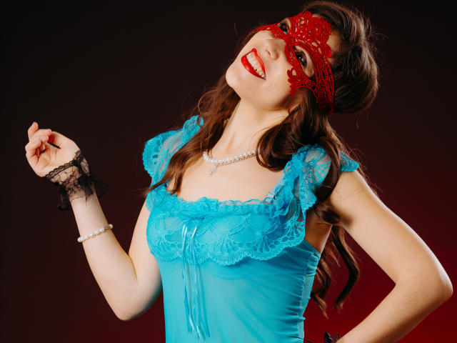 LadyXwinter mit roter Spitzenmaske, blauem Spitzentop und schwarzem Armreif, lächelnd, mit hochgehobener Hand, die ihre Brüste zeigt, vor dunklem Hintergrund..