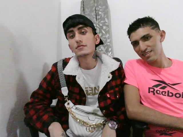 JuanAndNico, com um boné preto, camisa xadrez vermelha e carteira de corrente, está sentado ao lado de JuanAndNico, que veste uma camisa rosa da Reebok, ambos com cabelo curto e tatuagens..