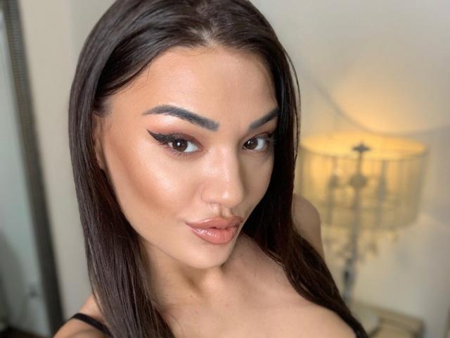 CataleyaLee has glossy brown lips, dark eyebrows, and long straight brown hair. She's wearing black with a nose ring and eyeliner that's winged. Her tits are partially visible.