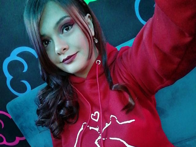 Katherin, con i capelli castano ricci, una felpa rossa e rossetto rosa, tiene in mano un selfie, mostrando il suo viso e la parte superiore del corpo, un piccolo piercing al naso e degli orecchini carini..