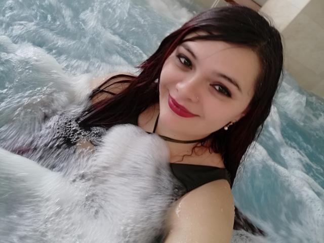 KatherinLatin with wet black hair, red lipstick, and black swimsuit, smiling in a bubbling water pool, holding foamy water in front of her tits.