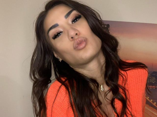 CataleyaLee with long dark hair, thick eyebrows, and full pout making a puffy kiss face, wearing an orange knit top, and a necklace, in a selfie.