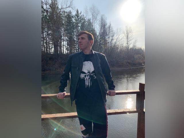 LarryHero staat bij een houten leuning, gekleed in een zwarte jas en shirt met een witte schedelprint, met zijn handen op de leuning, zonlicht achter hem, een bebost landschap op de achtergrond, water dat het licht reflecteert..