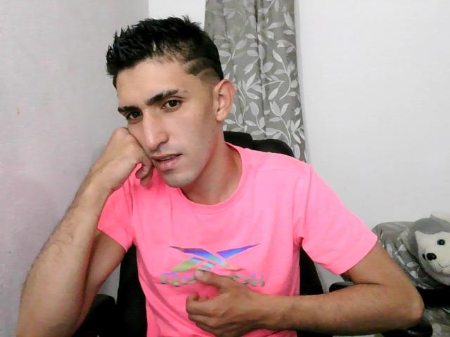 JuanAndNico, cabello corto y oscuro, vistiendo una camiseta rosa con un diseño, con el mentón apoyado en la mano, con una expresión ligeramente confundida, sentado en una silla, con la mano derecha sobre el estómago..