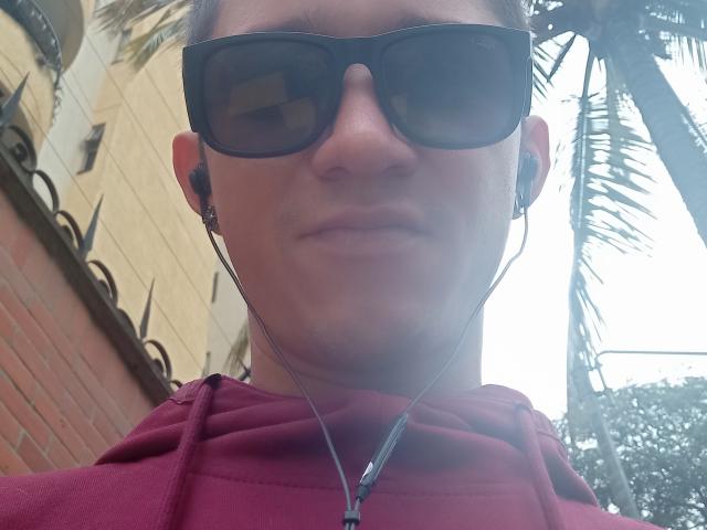 AhrilbLigan, con gafas de sol, auriculares y una sudadera roja, mirando ligeramente hacia abajo con una expresión neutra, cabello castaño corto y un ligero bigote..