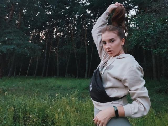 MissDi, con i capelli castani raccolti in uno chignon, indossa una felpa beige e un reggiseno sportivo nero, è in piedi in una foresta erbosa, guarda la fotocamera e ha una mano tra i capelli..