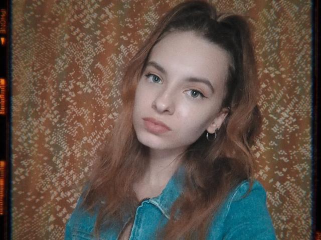 MissDi mit welligem, rotem Haar, die ein blaues Jeanshemd trägt, kleine Reifenohrringe und einen leichten Schmollmund, blickt mit grünen Augen aus dem Bild. Ihre Brüste sind nicht sichtbar..