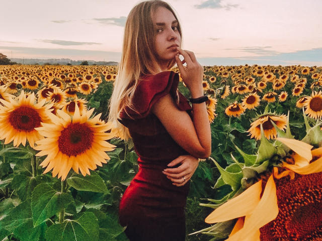 MissDi, com longos cabelos loiros, vestindo um vestido vermelho, em um campo de girassóis, mordendo o dedo, mostrando o bumbum, olhando por cima do ombro, céu ao pôr do sol, nublado..
