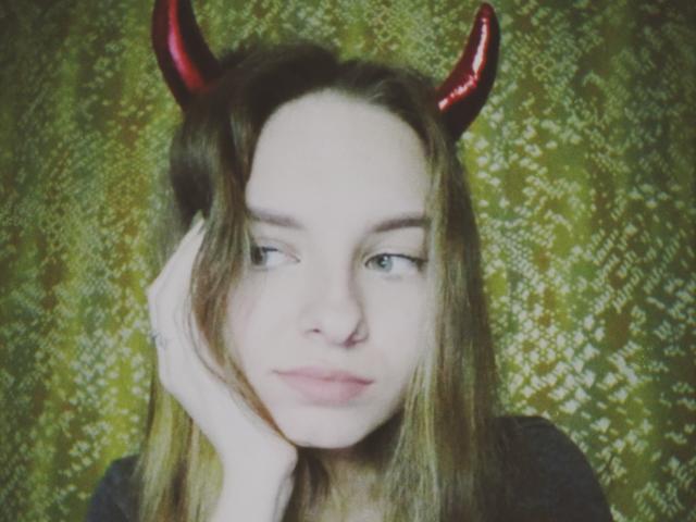 MissDi con corna da diavolo rosse, pelle pallida, occhi azzurri e capelli castani, appoggia il mento sulla mano, appare leggermente annoiata, indossa una maglietta nera..