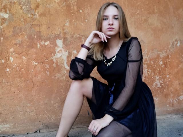 MissDi, con cabello rubio, vestido negro transparente, collar dorado y pulsera morada, está sentada con una rodilla levantada, mano en la barbilla, con expresión seria, mostrando su trasero y pechos pequeños..