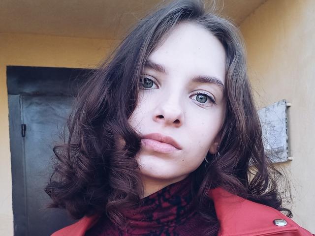 MissDi ha i capelli castano ondulati, la pelle chiara, gli occhi verdi e le labbra piene con un leggero sorriso. Indossa una giacca rossa sopra una maglia a collo alto scura..