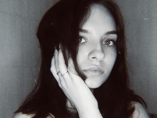 MissDi, com cabelo escuro, pele clara e olhos sérios, tocando o rosto com uma mão adornada com anéis, olhando diretamente para a câmera, em uma fotografia em preto e branco, ligeiramente texturizada..