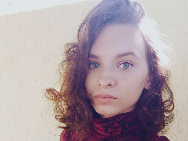 A MissDi tem olhos azuis, cabelo castanho ondulado e um cachecol vermelho com padrão, com uma expressão séria e um leve sorriso. Seu rosto está centralizado, com pele clara e iluminação natural suave..