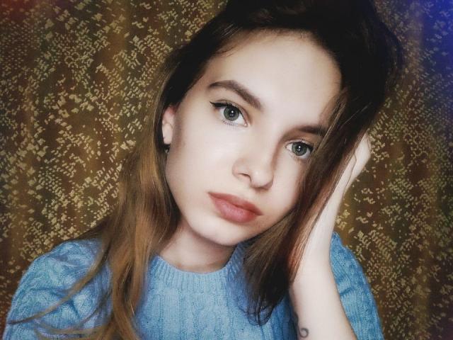 Miss Di mit hellgrünen Augen, vollen, rosafarbenen Lippen und einem blauen Pullover, hat braunes Haar, ein Tattoo am rechten Handgelenk und einen ernsten Gesichtsausdruck, wobei sie mit ihrer linken Hand ihr Haar berührt..