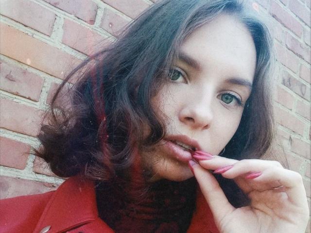 MissDi, avec ses cheveux bruns ondulés, ses yeux verts et ses lèvres roses, mordant son ongle peint en rose, portant un manteau rouge, regardant légèrement sur le côté, sur une photo de profil..