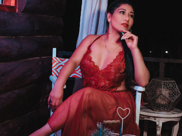 KhloeLawson with long black hair, red lace lingerie, and sheer red skirt, sitting on a chair, finger to mouth, looking to the side, with a heart pattern on skirt.