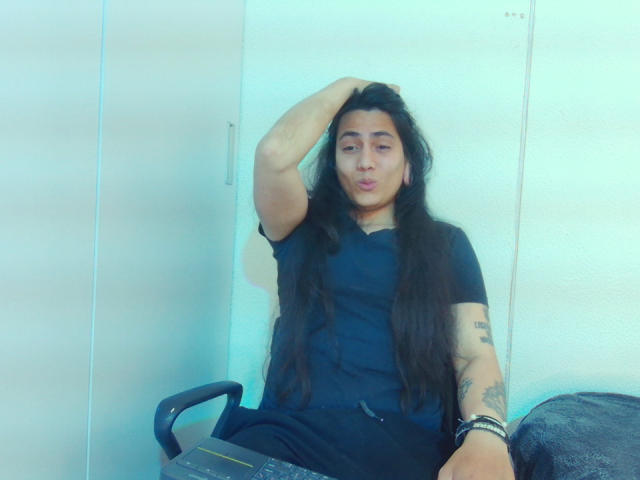 Een persoon genaamd Igorendon, met lang zwart haar, een zwart shirt en tatoeages, die een zuur gezicht trekt met één arm achter het hoofd, zittend op een stoel, waarbij de blote borst en zwarte broek te zien zijn..