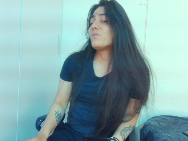 Igorendon, con lunghi capelli neri, indossa una maglietta nera, ha le labbra tumefatte, tatuaggi sulle braccia, è seduto su una sedia, foto con tonalità blu, indossa un braccialetto nero..