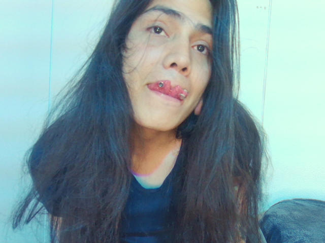 Igorendon, com cabelos longos e castanho-escuros, levemente desalinhados, tem um piercing rosa no lábio inferior e está vestindo uma blusa preta, olhando para a câmera com um sorriso discreto..
