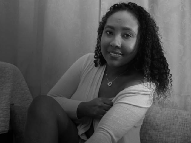 CaroldelaTorre with curly hair, wearing a light cardigan, smiling, sitting on a couch, hand on chest, showing tits, necklace with heart pendant, black and white photo.