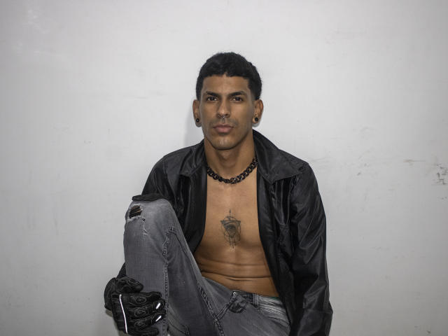 MarcoCarter, sem camisa, jaqueta de couro preta, calças jeans cinzas rasgadas, luvas pretas, colar de corrente, tatuagem no peito, cabelo curto e cacheado preto, piercing no nariz, protetores auriculares, sentado, expressão neutra, parede cinza..