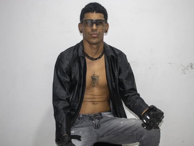 MarcoCarter, shirtless, wearing black jacket, glasses, black bead necklace, tattoo on chest, grey ripped jeans, black gloves, short black hair, white wall background, looking at camera.