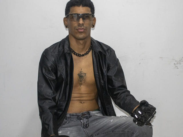 Marco Carter, con i capelli neri corti, che indossa occhiali trasparenti, una collana a catena nera, una camicia nera aperta, un tatuaggio sul petto, guanti neri e jeans strappati, seduto di fronte a un muro bianco liscio..