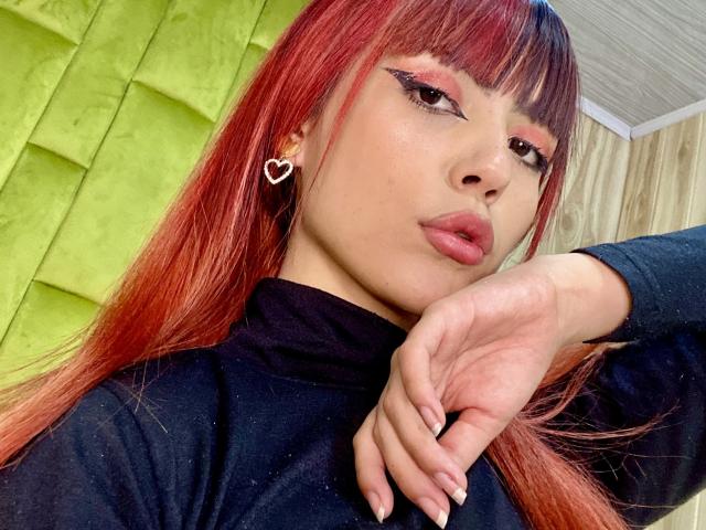 MagicianFoster, con cabello rojo, aretes en forma de corazón, un cuello alto negro, delineador de ojos rosa y labios carnosos, con el mentón apoyado en la mano, luciendo seductora. Los senos no son visibles..
