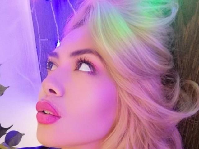 SelleniaX mit blonden, regenbogenfarbenen Haaren, die nach oben schauen, mit pinkem Lippenstift und Eyeliner, hat ein sinnliches, attraktives Gesicht mit einem leichten Rouge, in einer farbenfrohen, neonbeleuchteten Umgebung..