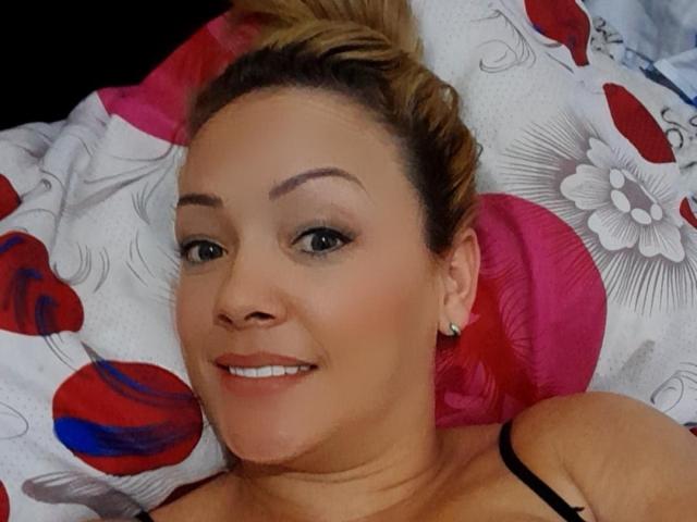 A DevoraQuiins tem cabelo loiro, olhos verdes e está sorrindo, com a cabeça apoiada em um travesseiro floral rosa e vermelho. Ela está vestindo uma alça preta, mostrando seus seios..