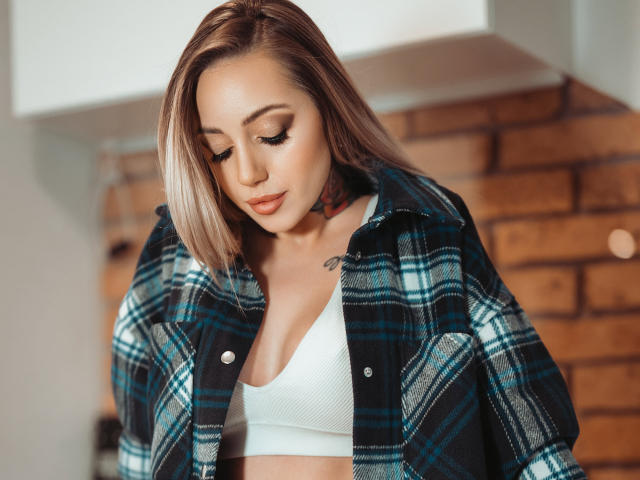 CoralieFlower with blonde hair, wearing a blue plaid shirt over a white bra, looking down with a slight smile, showing her tits. Soft makeup, gentle expression.