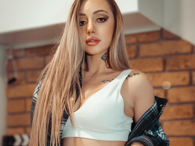 CoralieFlower with long blonde hair, white sports bra, and unbuttoned denim jacket, showing small tattoos on her neck and shoulder, with her tits slightly visible.