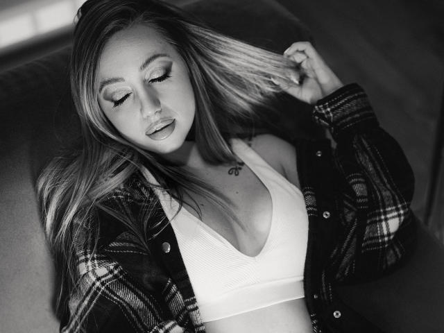 Black-and-white photo of CoralieFlower with long blonde hair, eyes closed, wearing a plaid shirt over a white sports bra, touching her hair, showing small tits and a tattoo.