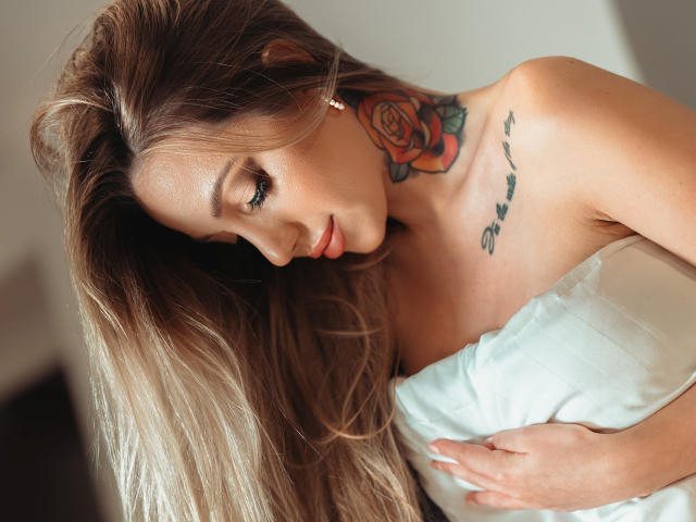 CoralieFlower with long blonde hair, tattooed rose on neck, and "stay" on chest, looking down, holding white pillow, topless, showing her tits, soft light, gentle expression.