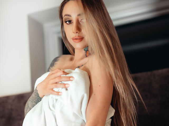 CoralieFlower with long blonde hair, winking, holding a white pillow over her tits, tattoo on right shoulder, tattoo on left neck, arm tattoo, sitting on brown couch.