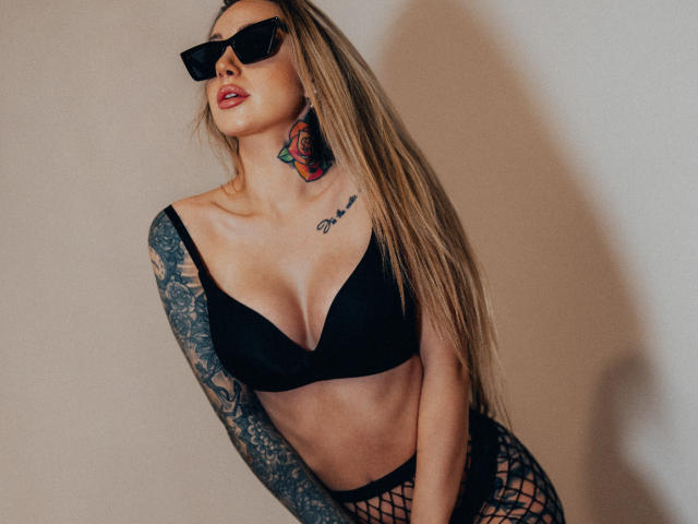 CoralieFlower, com seus longos cabelos loiros, sutiã preto, calças de rede, tatuagens e óculos de sol pretos, exibindo seus seios e braço tatuado, aparentando ser sexy e confiante..