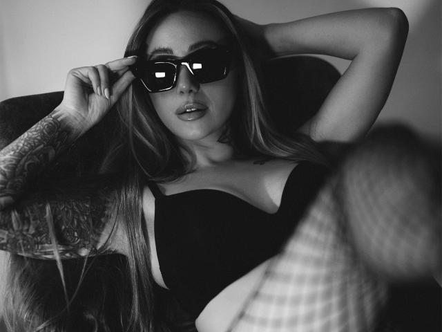 Fotografía en blanco y negro de CoralieFlower con cabello largo, tatuajes en el brazo derecho, usando gafas de sol oscuras, un top negro sin tirantes, bajándose las gafas de sol de forma seductora con la mano derecha, labios ligeramente entreabiertos..