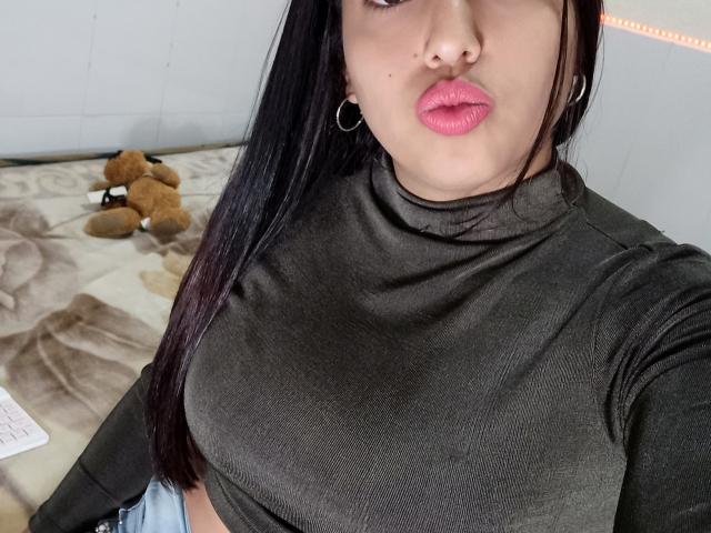 SamiLove with pouty pink lips, dark brown long hair, hoop earrings, green turtleneck, showing her boobs, laying on bed with brown teddy bear.