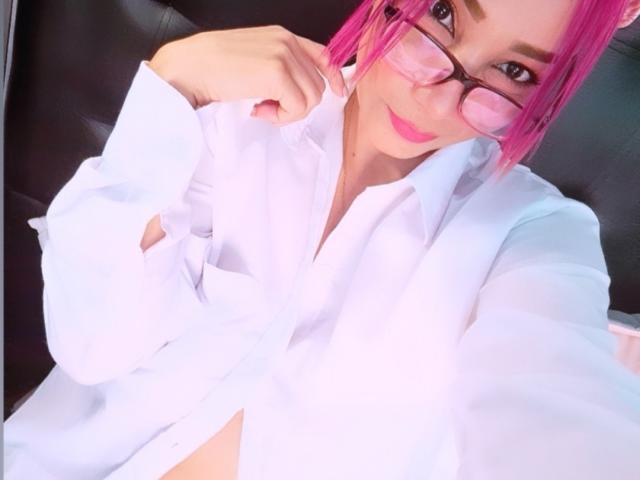 AmberDemons with pink hair, glasses, and white shirt, holding collar, showing bare midriff, smiling, upper body, selfie, pink lipstick, black leather chair.