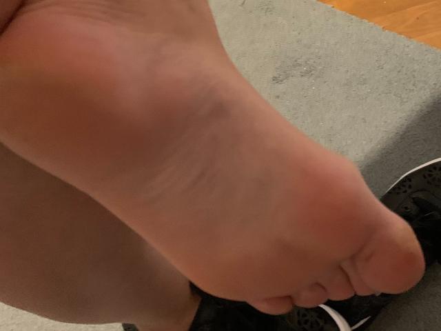 QueenKaly's bare foot with a slightly pink sole, her toes pointed downward, and a black shoe with white stripes partially visible in the bottom right.