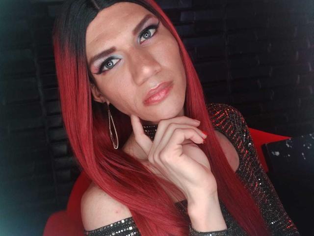 LunaGransTs with red hair, blue eyes, and glossy pink lips, wearing a black sequined top, gold hoop earrings, and touching her chin, looking directly at the camera.
