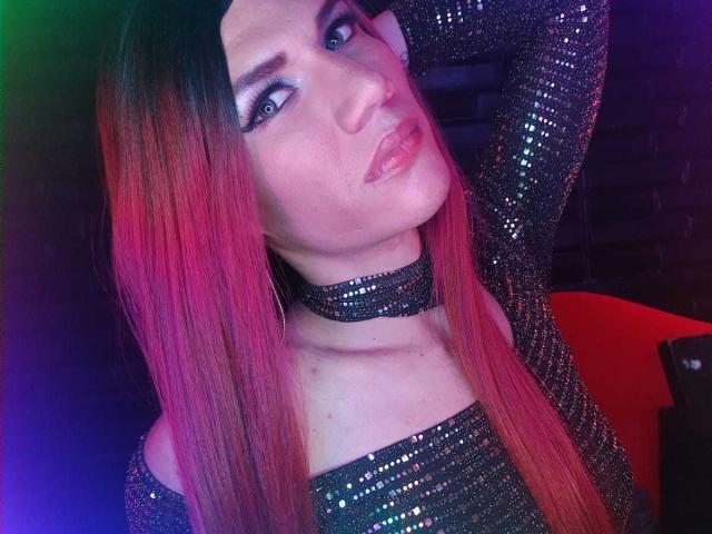LunaGransTs with pink and black ombre hair, wearing sparkly black top and choker, blue eyes, pink lipstick, looking up with hand in hair, in colorful light.