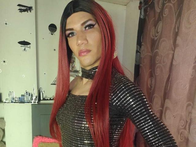 LunaGransTs with long red hair, wearing a shiny black dress, has a serious expression, visible makeup, and a choker, standing in a room with pink curtains.