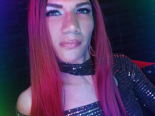 LunaGransTs with long pink-red hair, blue eyes, and glossy pink lips, wearing a sparkly black choker and dress, looking up with a slight smile. Boobs are partially visible.