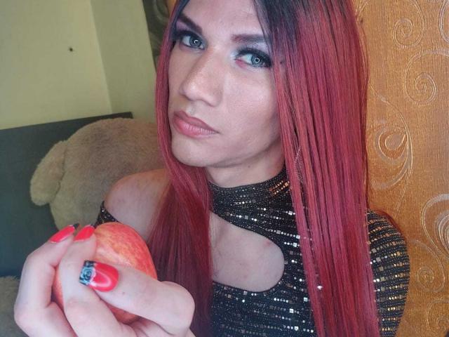 LunaGransTs con cabello rojo largo, ojos azules y lápiz labial rosa, sosteniendo una manzana roja, vistiendo una blusa negra con lentejuelas, con uñas rojas y un anillo azul, con una expresión seria..