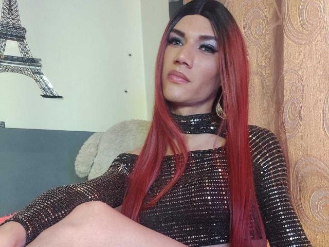 LunaGransTs with red and black ombre hair, wearing a sparkly black top, sitting with legs crossed, looking up with a slight smile, wearing gold earrings.