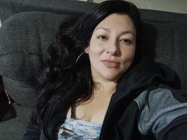 SakotoDika with black wavy hair, medium tits, wearing a gray hoodie and hoop earrings, lying on a dark gray couch, smiling with pink lipstick.
