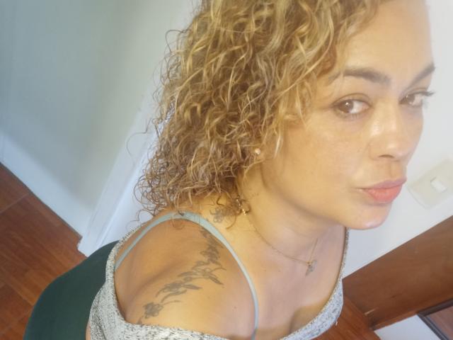SolmaryHot has curly blonde hair, light brown skin, and a tattoo on her right shoulder. She's wearing a sparkly top, showing her small tits and a green bottom.