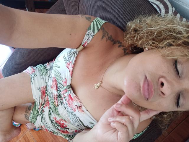 SolmaryHot with curly blonde hair, tan skin, and a floral dress, lying on a brown couch, pouting with eyes closed, hand near face, gold necklace, and tattoos visible.