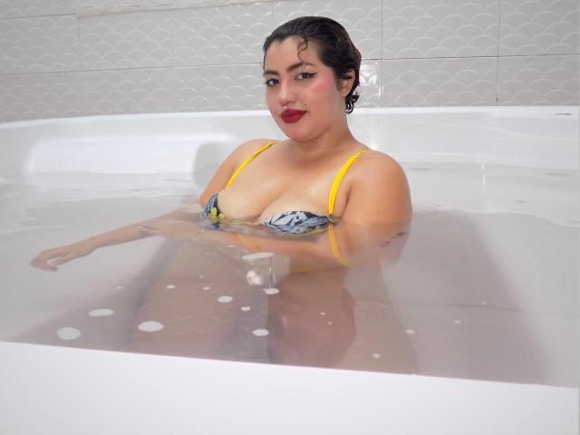 CharlizeAddams dans une baignoire, l'eau lui arrivant à la poitrine, portant un haut de maillot de bain à motifs jaunes et bleus, montrant ses seins, cheveux foncés et mouillés, rouge à lèvres rouge, souriante..
